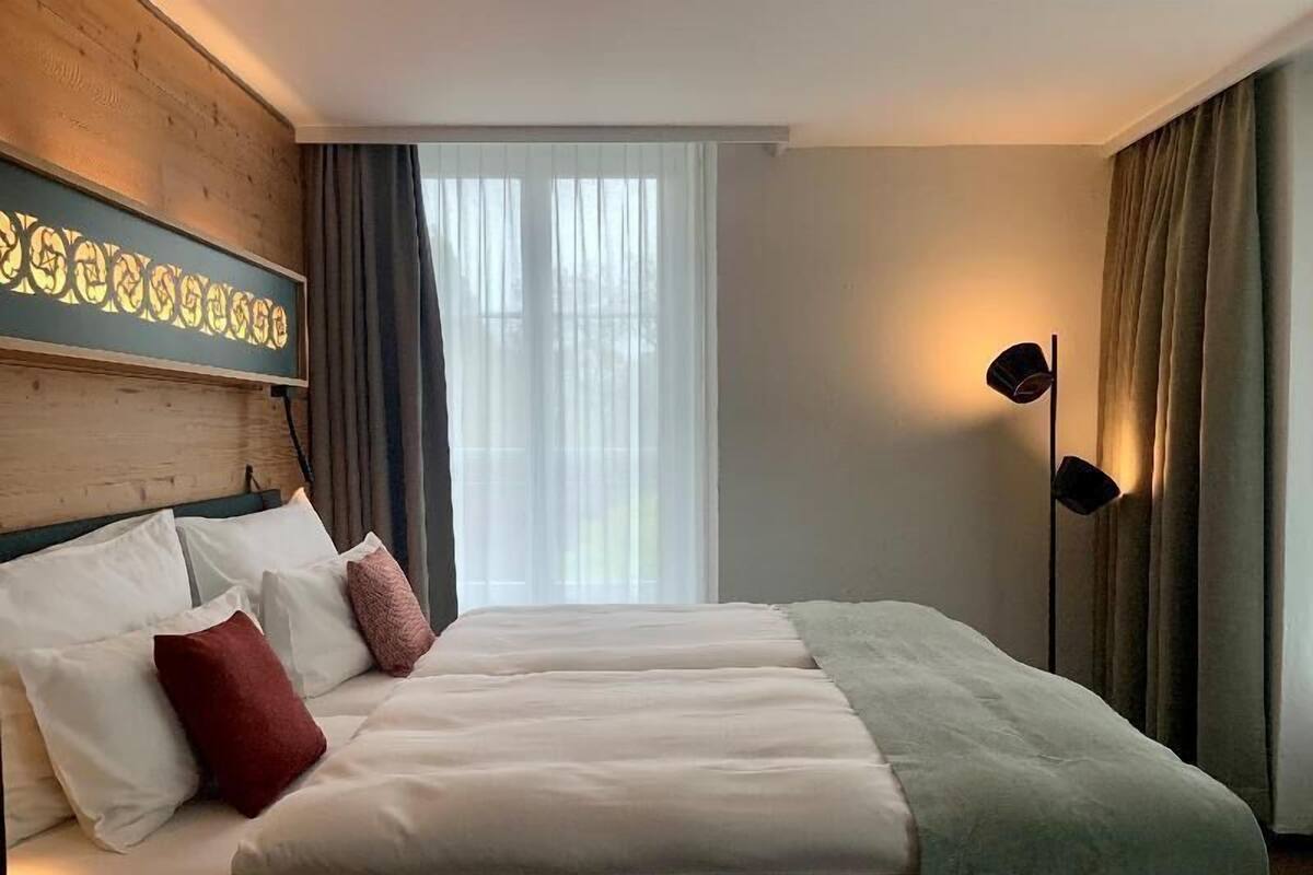 A hotel room with a large bed, a headboard with a decorative panel, a floor lamp, and a window with curtains.