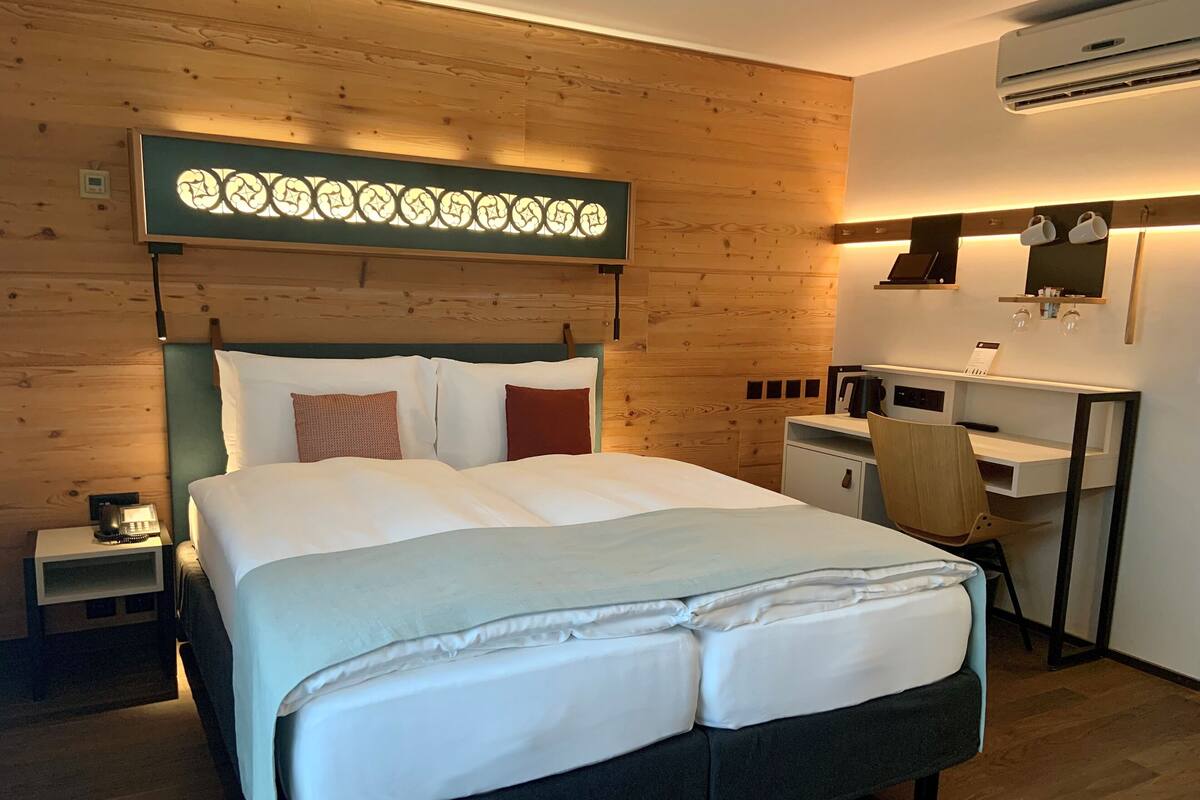 A hotel room with a large bed, a desk, a chair, a small cabinet, and an air conditioning unit.
