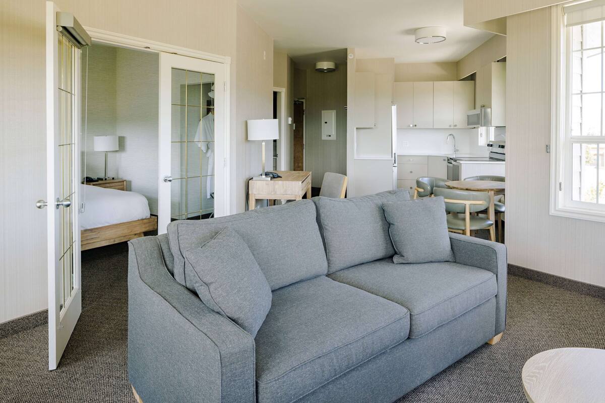 A modern hotel room with a grey sofa, a small dining table, a bed with a wooden headboard, a desk with a lamp, and a kitchenette in the background.