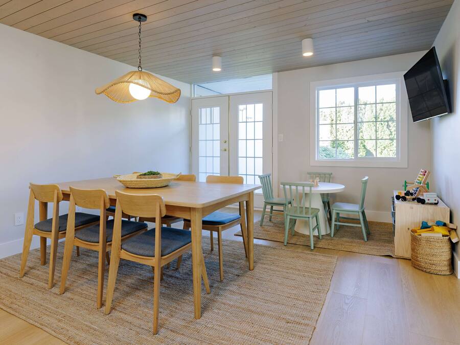 A dining area with a wooden table and chairs, a hanging light fixture, a television mounted on the wall, and a window with a view of trees.