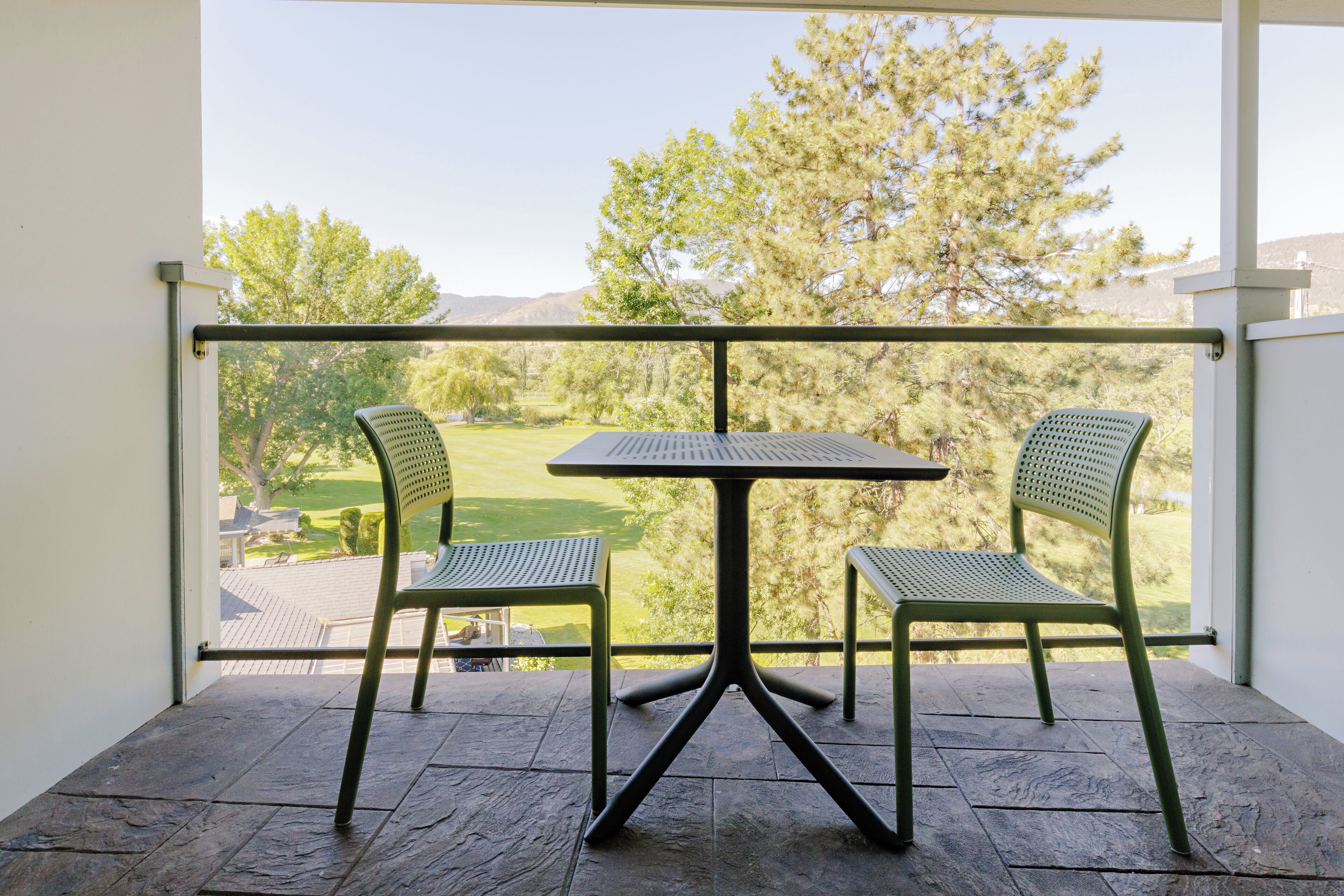 A balcony with a table and two chairs, overlooking a green landscape with trees and a distant view of a house.
