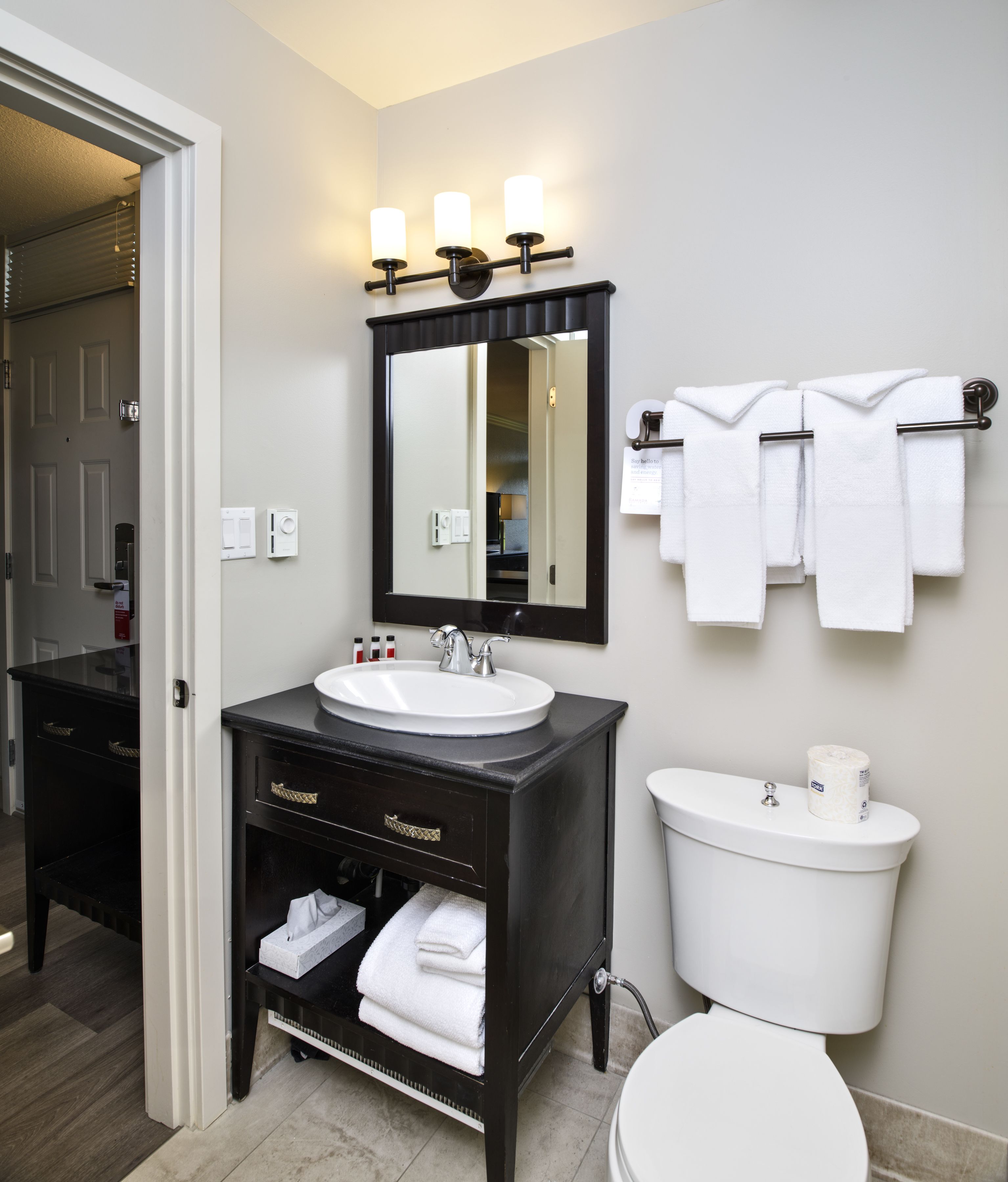 A bathroom with a black vanity, white sink, and a toilet. There are three towels hanging on a towel rack, and a mirror above the sink.