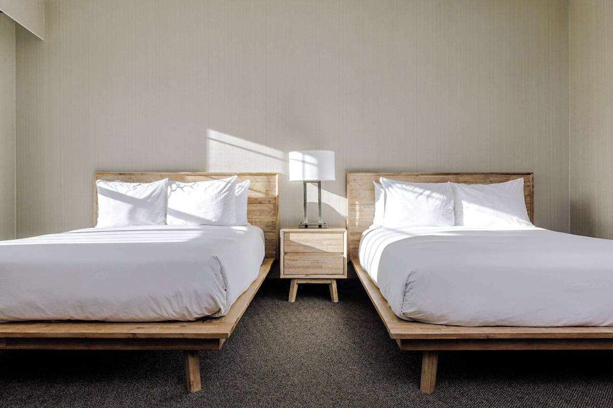 A hotel room with two wooden beds, white bedding, a small bedside table with a lamp, and a carpeted floor.