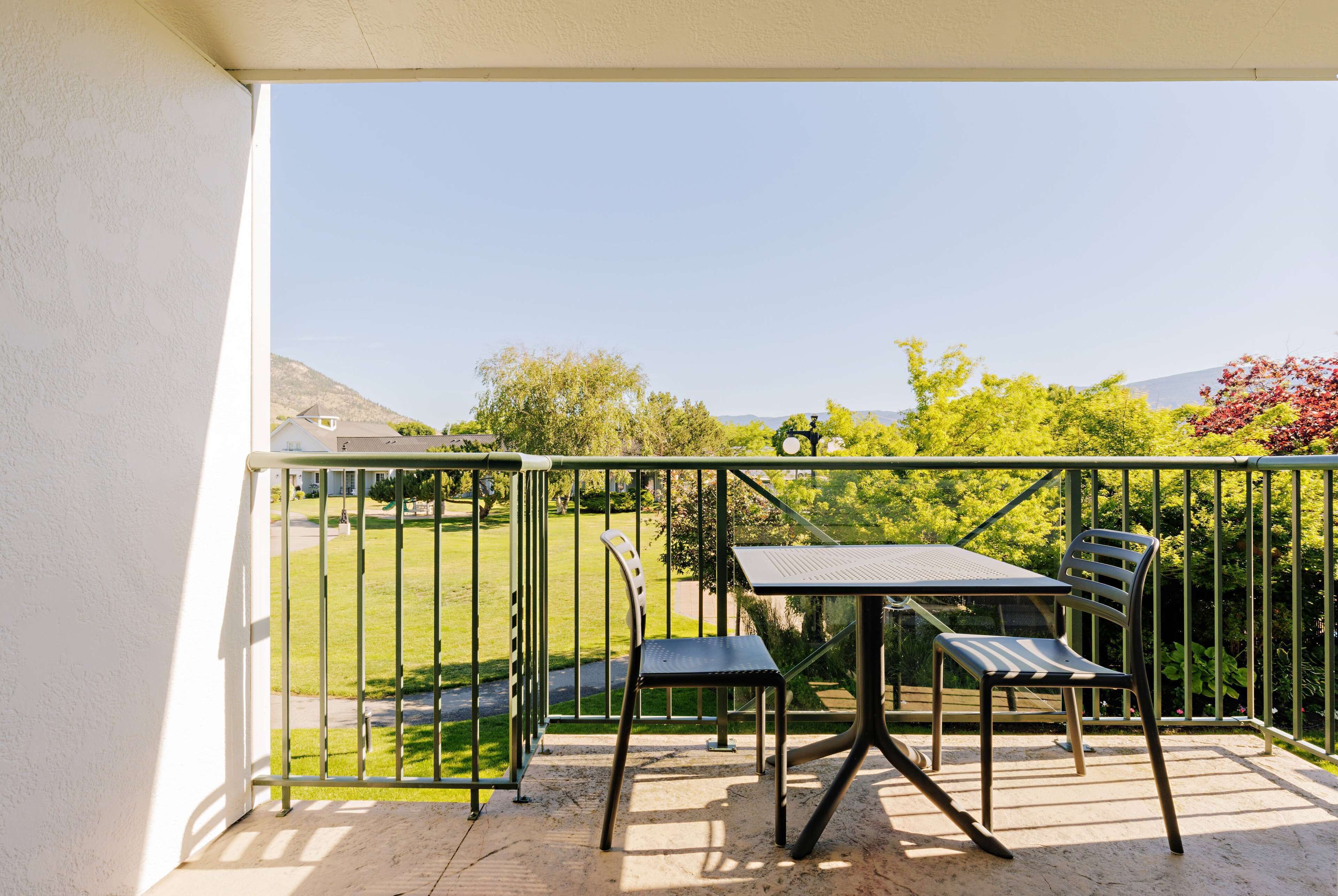 Un balcone con tavolo e due sedie, ringhiera in metallo e vista su un prato verde e alberi.