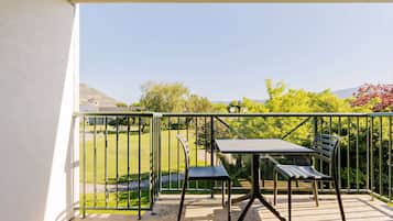 A balcony with a table and two chairs, a metal railing, and a view of a green lawn and trees.
