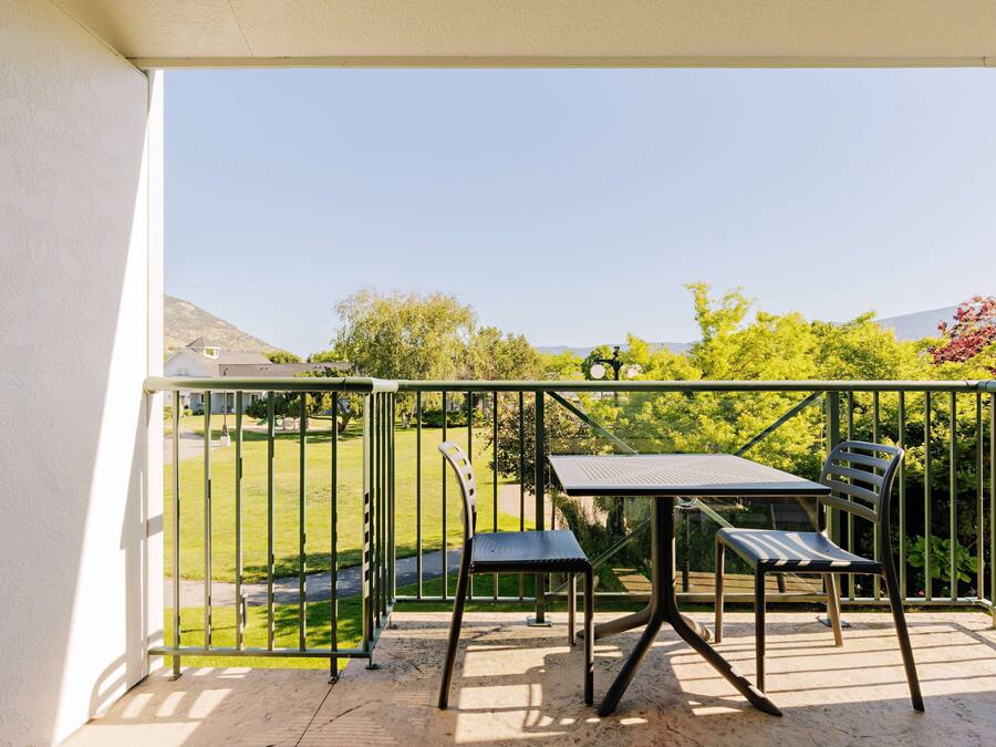 A balcony with a table and two chairs, a metal railing, and a view of a green lawn and trees.