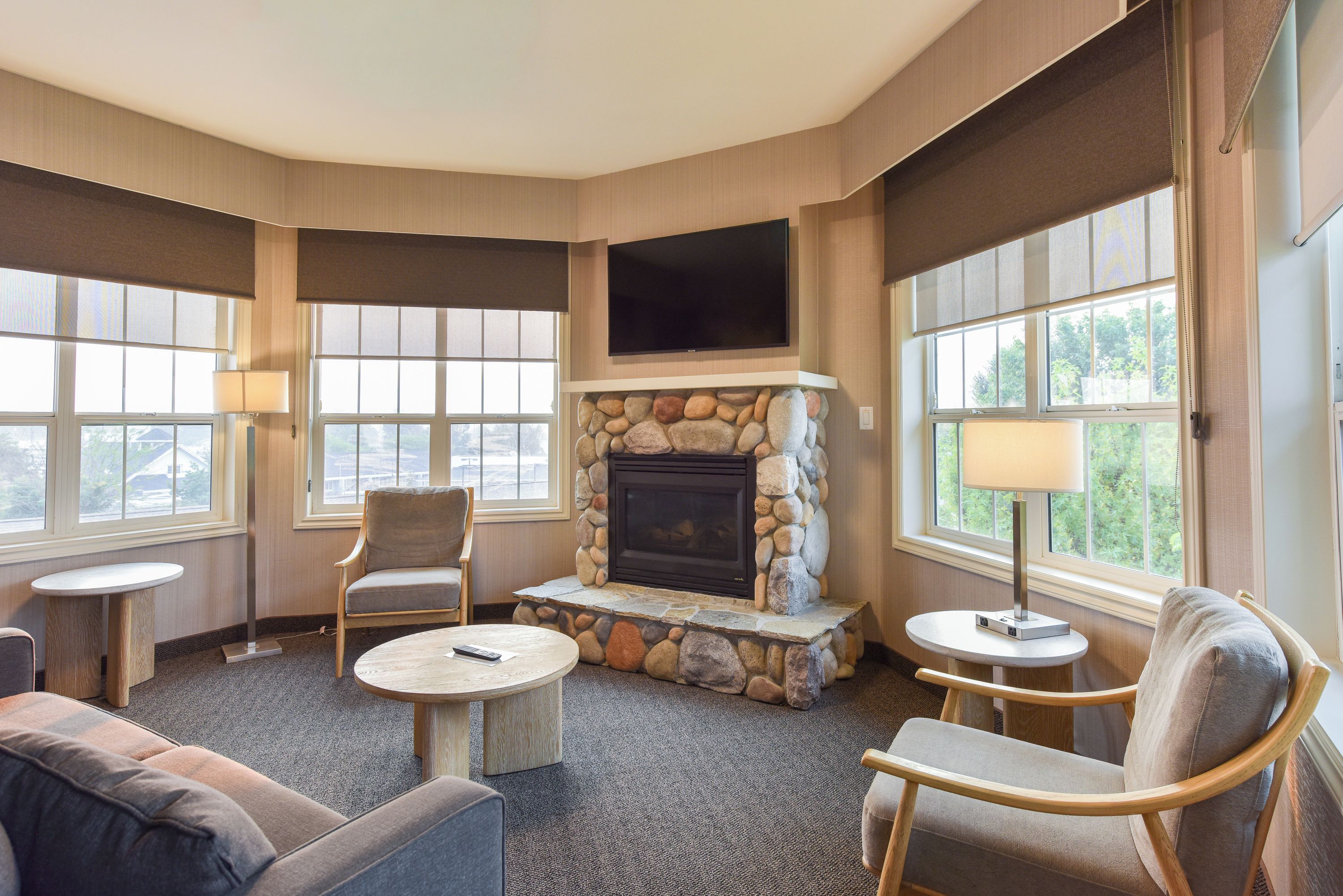 A hotel room with a stone fireplace, two armchairs, a sofa, a flat-screen TV, and large windows.