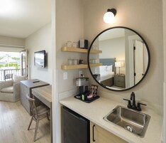 Una habitación de hotel compacta con kitchenette, una cama con cabecera, un espejo redondo y una pequeña zona de estar con una silla y una mesa.