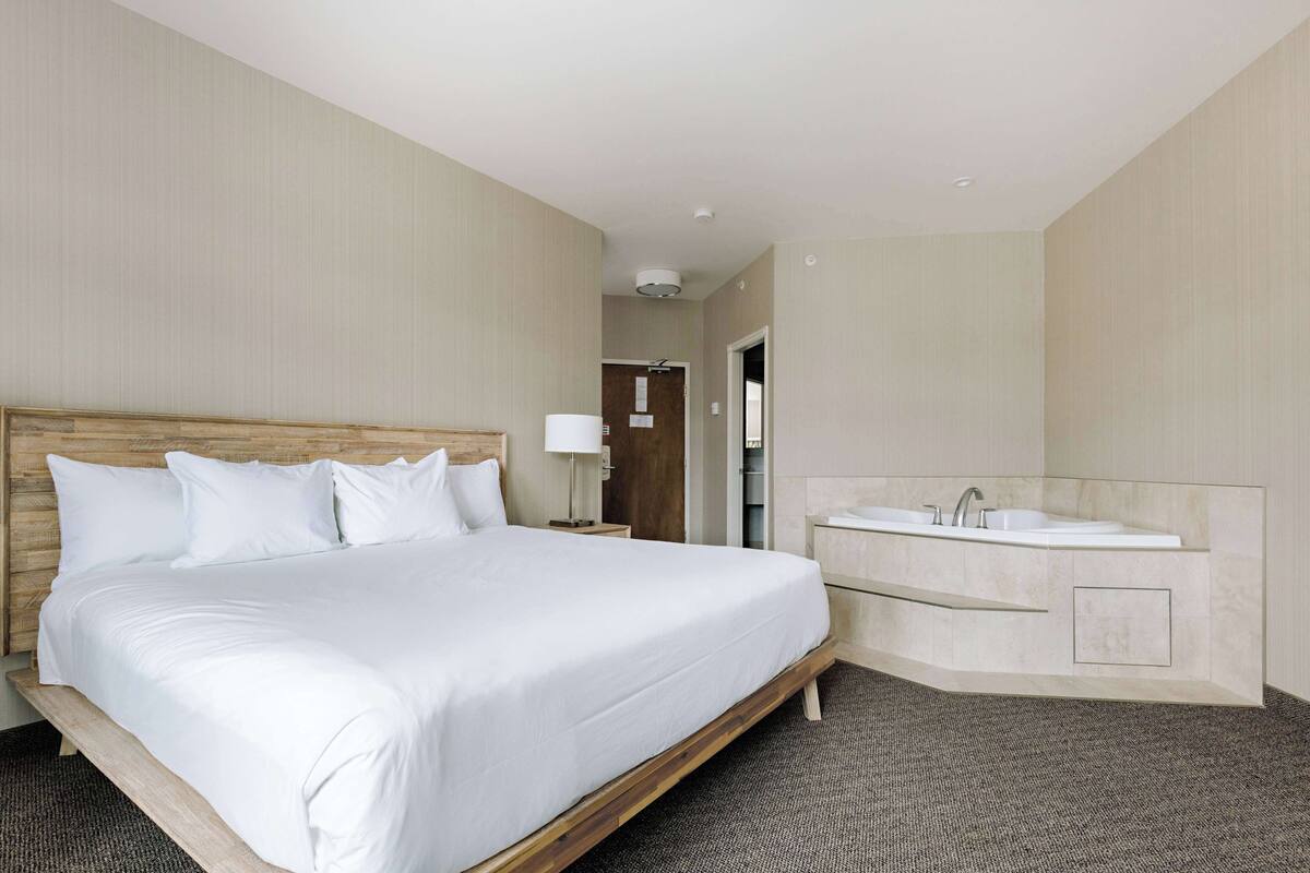 A hotel room with a large bed, a wooden headboard, a bedside table with a lamp, and a modern bathroom with a glass-enclosed sink and mirror.