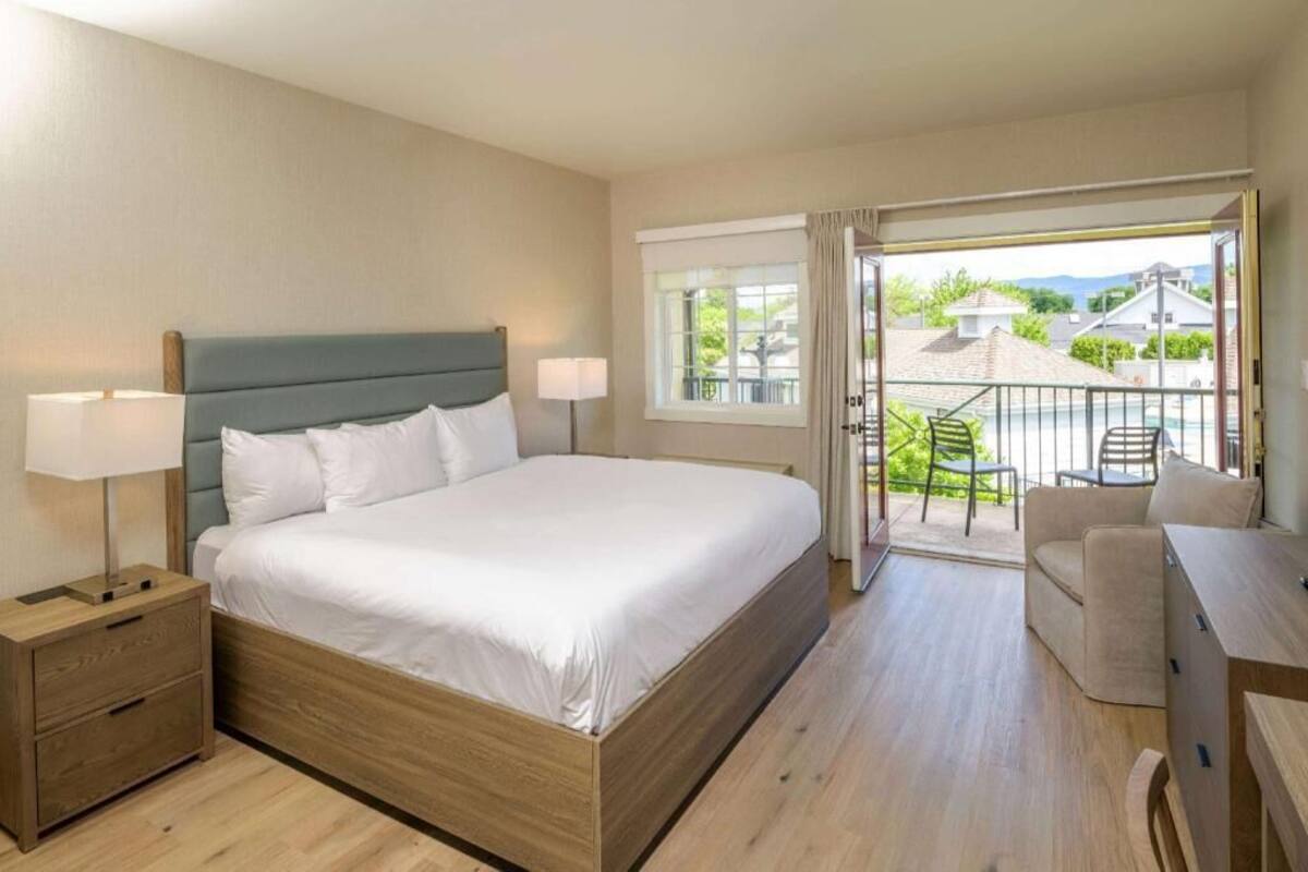 A hotel room with a bed, bedside lamps, a wooden nightstand, a balcony with a chair, and a view of the outside.