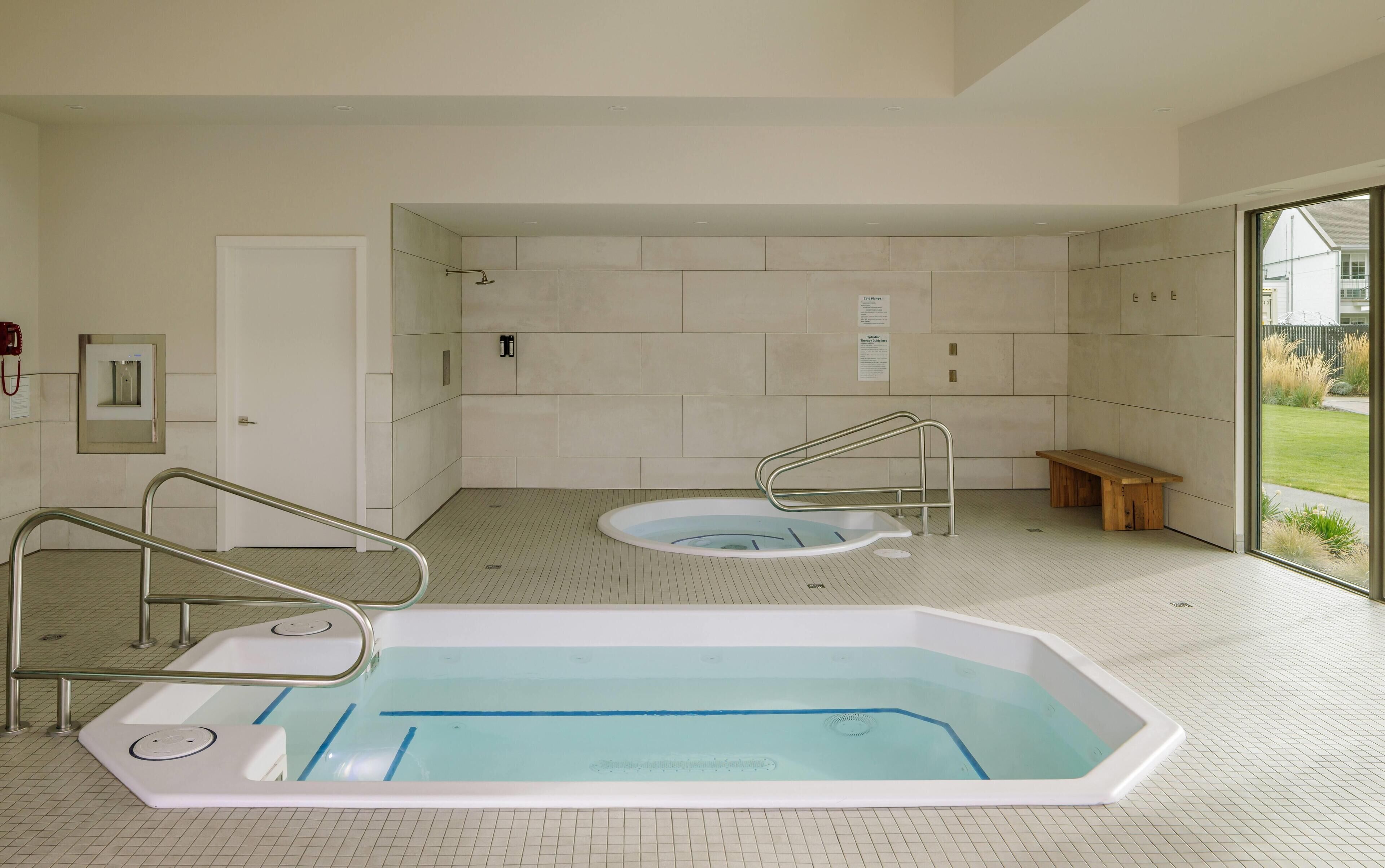 Een modern binnenzwembad met een bankje en een jacuzzi, omgeven door betegelde muren en een groot raam.
