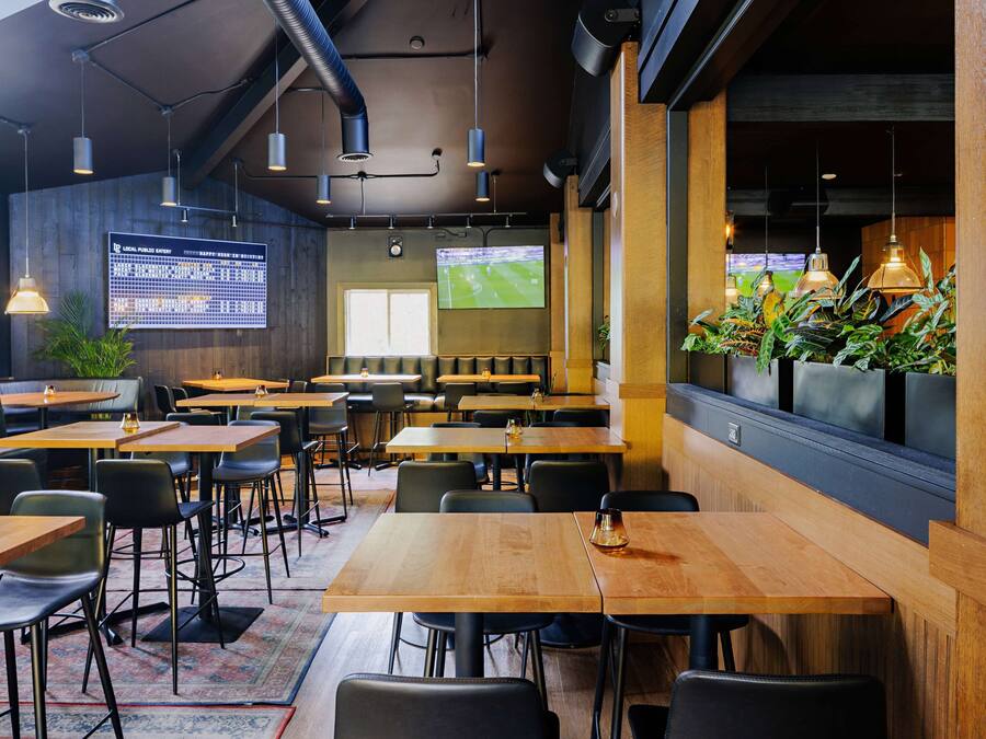 A restaurant with wooden tables and chairs, a large digital screen, and a TV displaying a sports event.