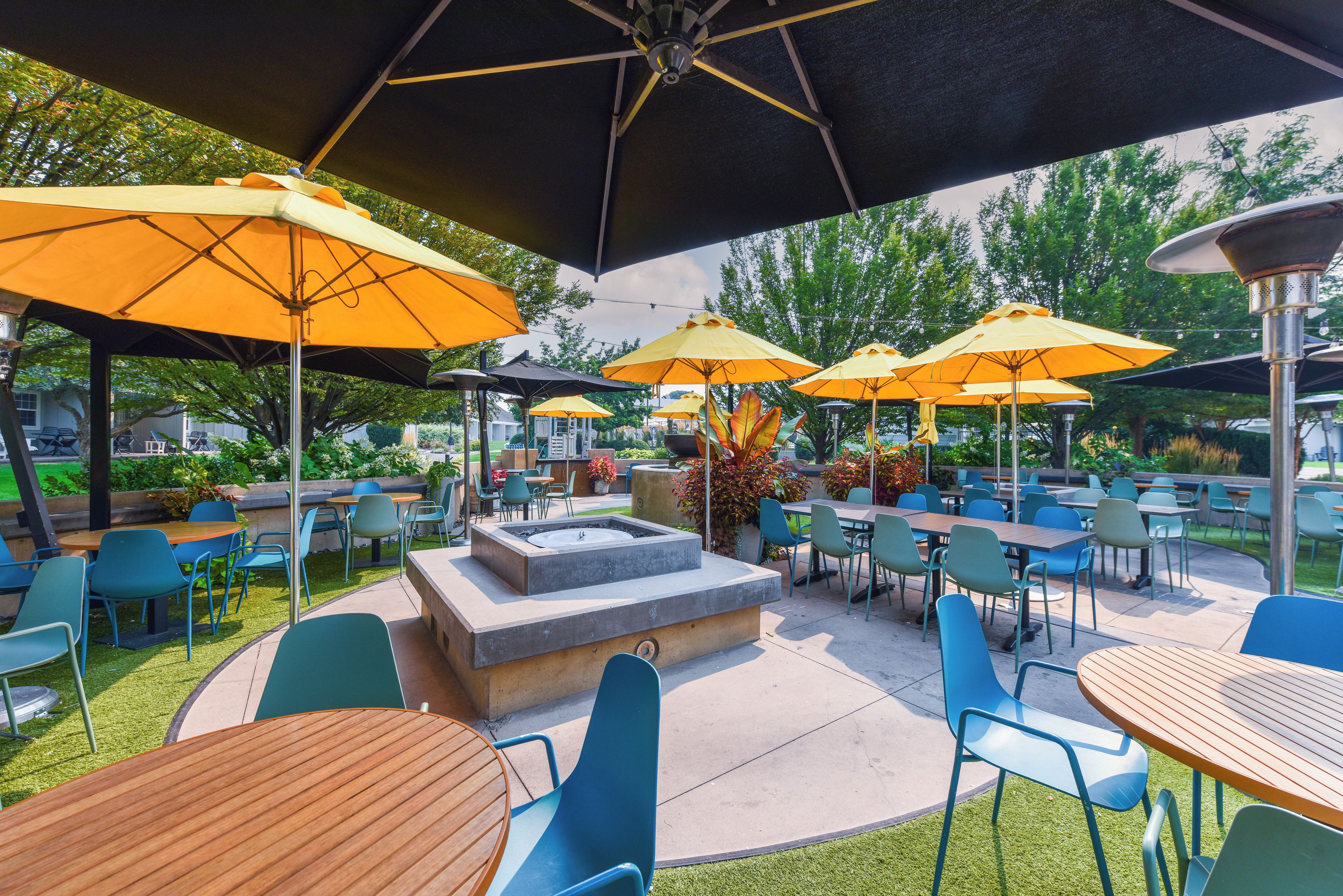 An outdoor patio area with multiple tables and chairs, large umbrellas, and a central fire feature.
