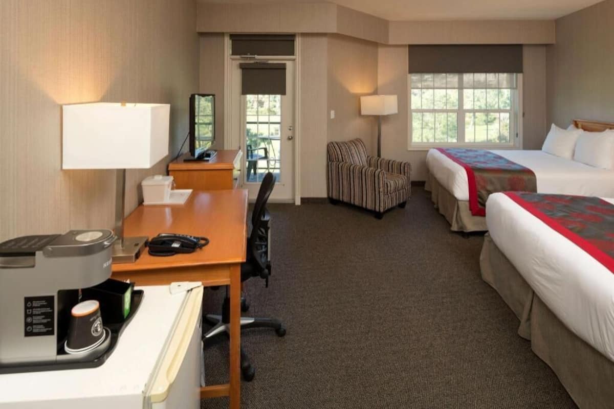A hotel room with a desk, two beds, a chair, a coffee maker, and a window.