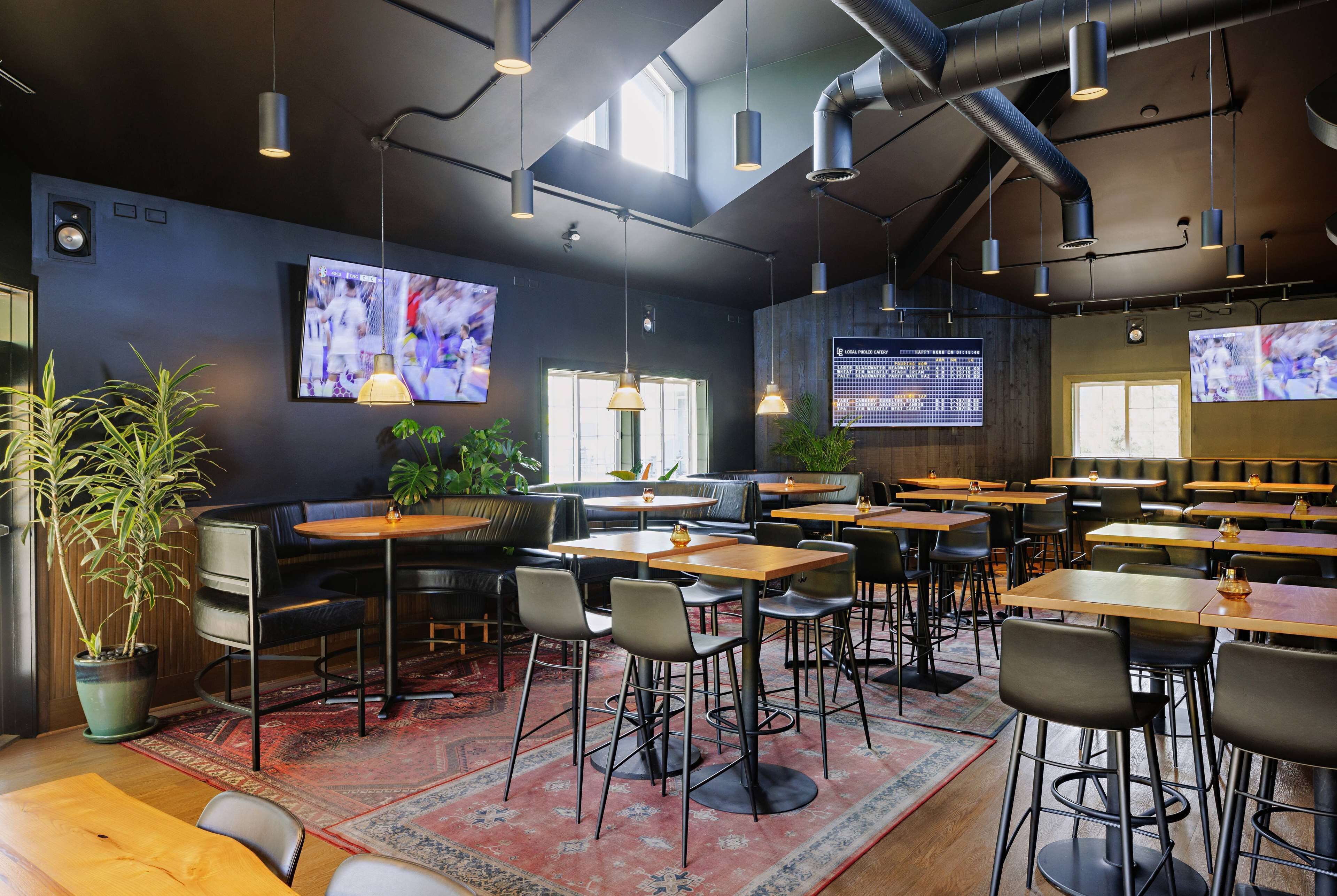 A modern restaurant with multiple tables and chairs, large screens displaying content, and a cozy atmosphere with plants and wooden accents.