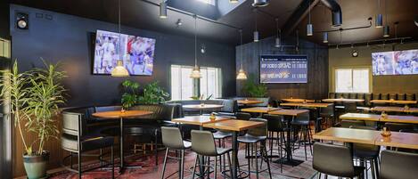 A modern restaurant with multiple tables and chairs, large screens displaying content, and a cozy atmosphere with plants and wooden accents.