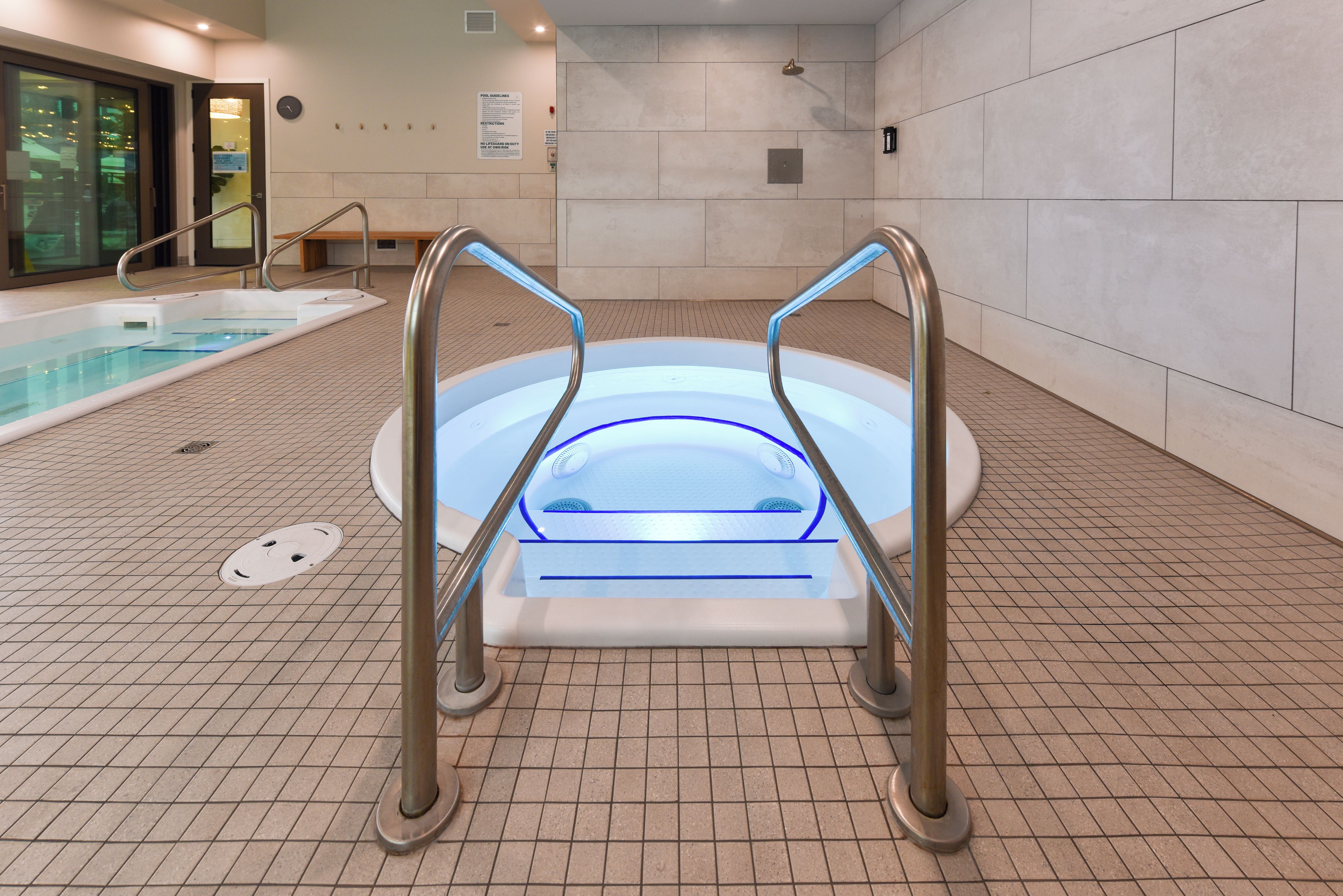 A hot tub with a glass enclosure and metal railings, surrounded by a tiled floor and a pool.