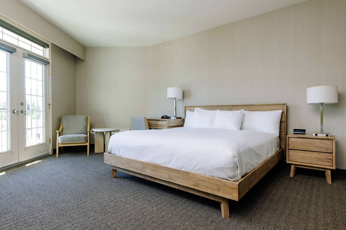 A hotel room with a large bed, two bedside tables, a chair, a small table, and a large window.