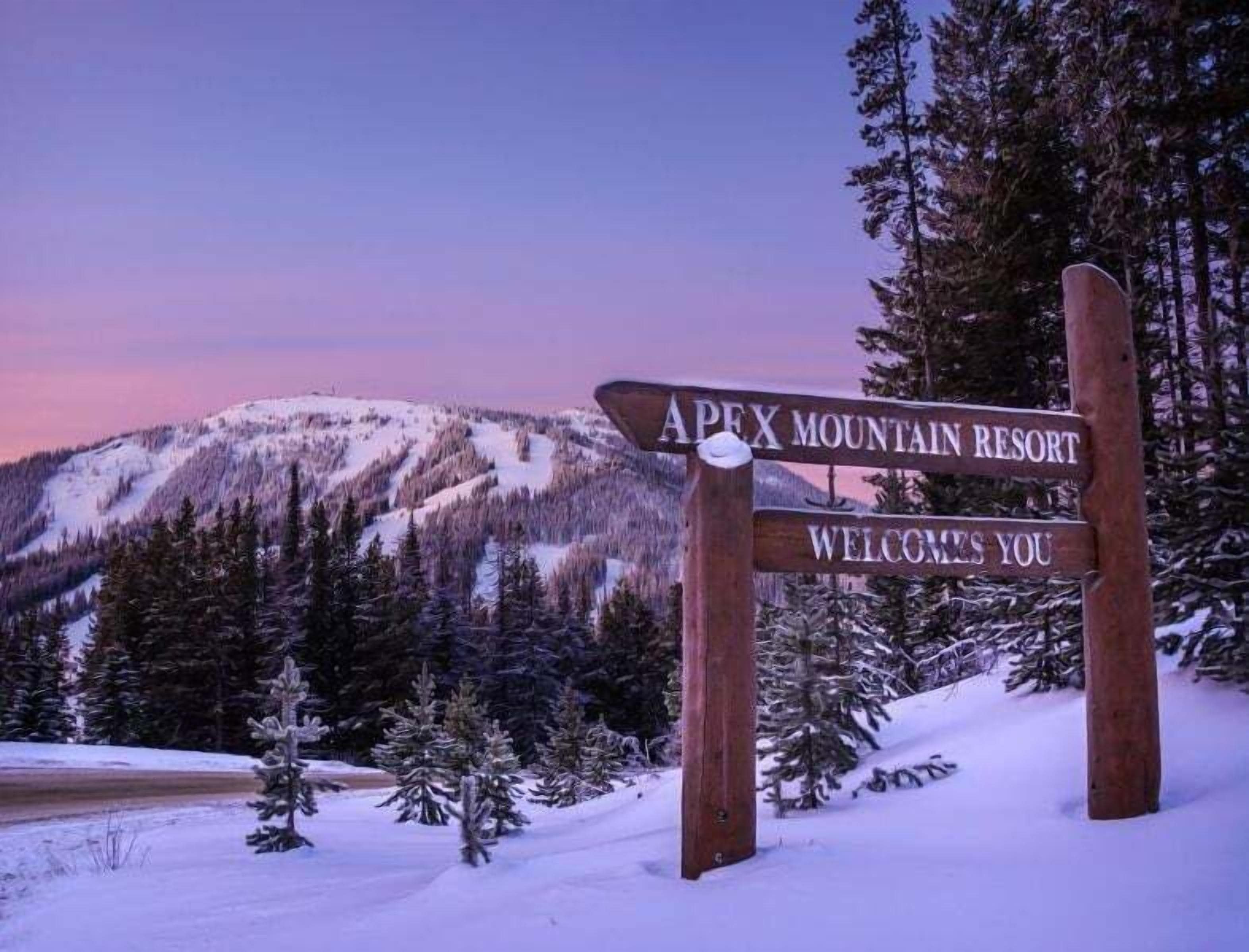 Un letrero de madera con el texto "Apex Mountain Resort" y "Bienvenido" sobre un fondo de árboles y montañas cubiertas de nieve.