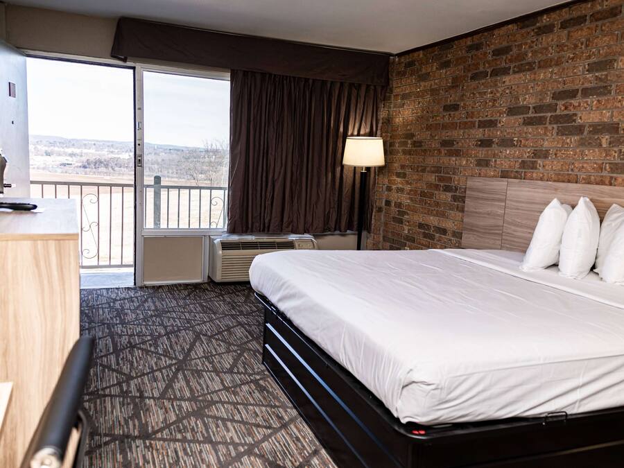 A hotel room with a large bed, a desk, a chair, a window with curtains, an air conditioning unit, and a brick wall.