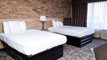 A hotel room with two beds, a brick wall, a large window with curtains, and a patterned carpet.