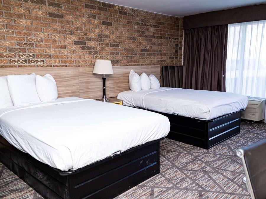 A hotel room with two beds, a brick wall, a large window with curtains, and a patterned carpet.