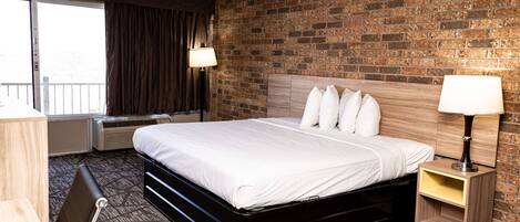 A hotel room with a brick wall, a bed with white bedding, a nightstand with a lamp, and a desk with a chair.