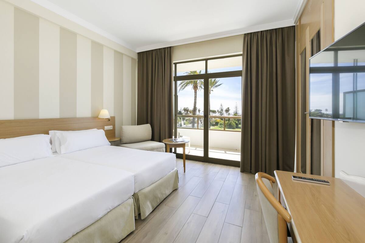 Habitación de hotel con una cama grande, un pequeño sofá, un escritorio y un balcón con cortinas.