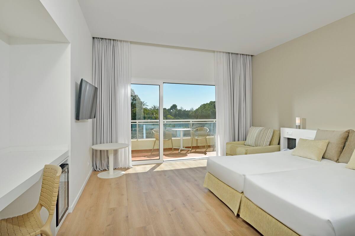 Habitación de hotel con una cama grande, televisión, balcón con mesa y sillas, y vista a zonas verdes.