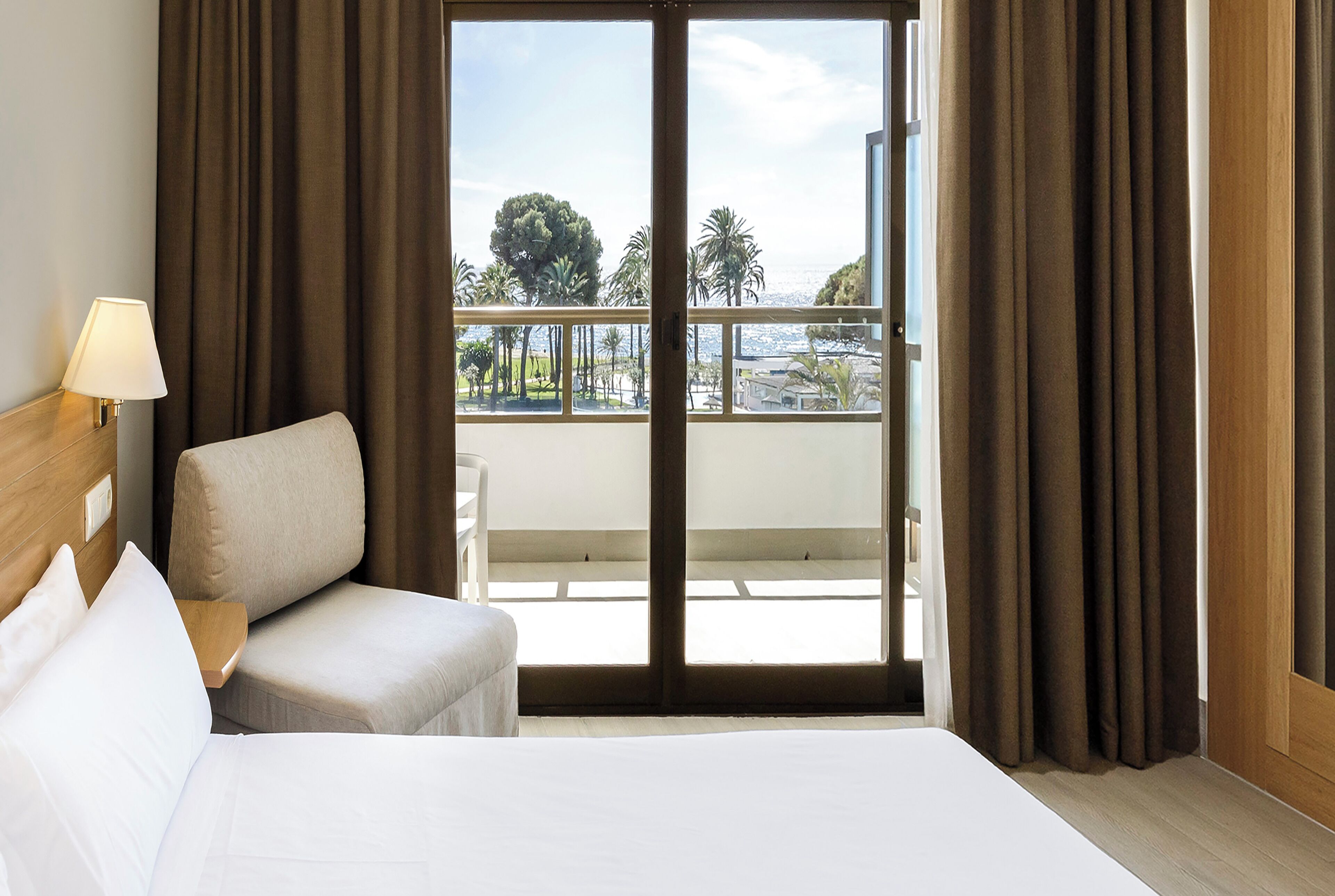 Habitación de hotel con una cama, una silla, una lámpara y vistas al exterior a través de una puerta corredera.