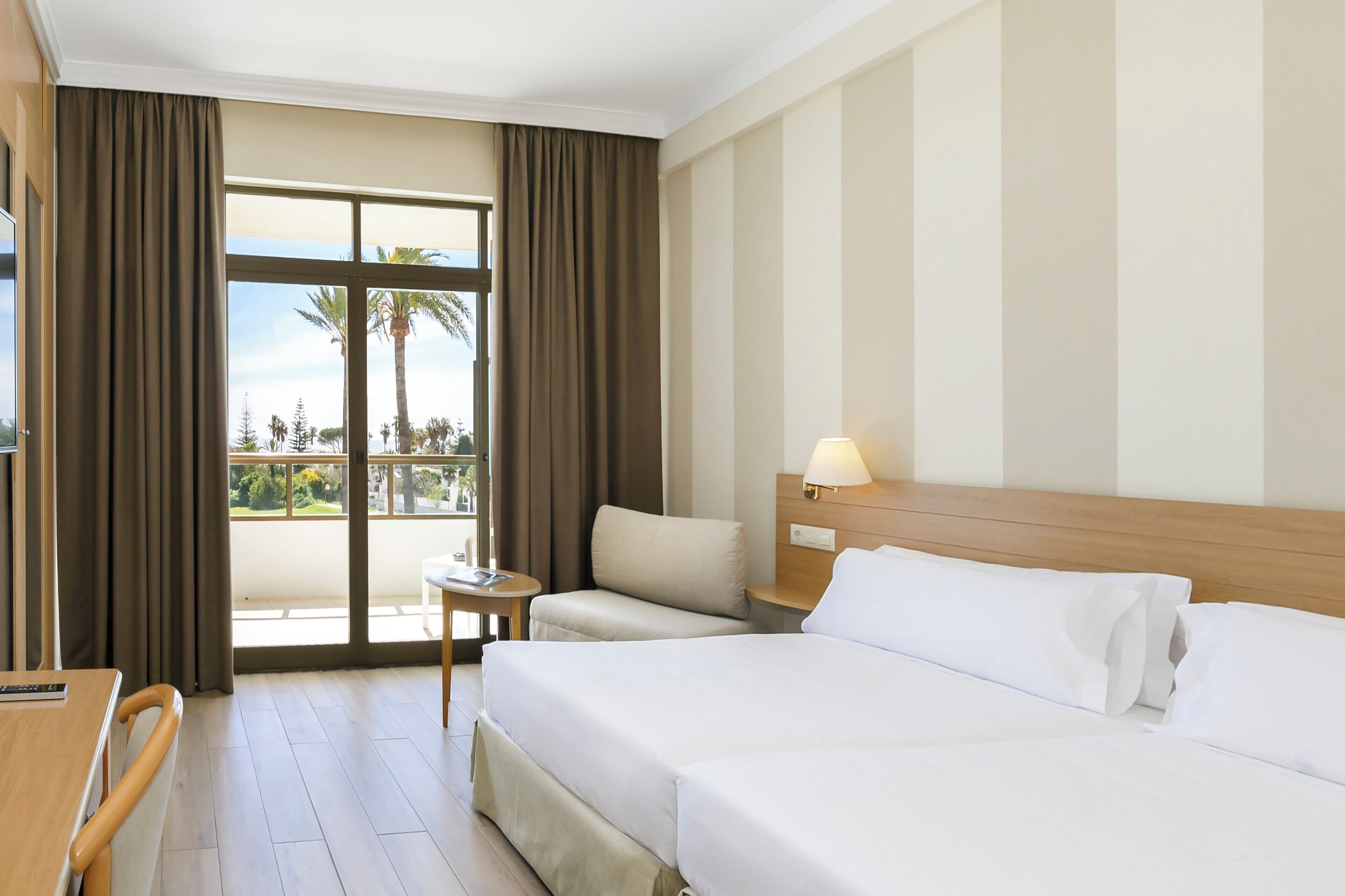 A hotel room with a large bed, a bedside table, a chair, and a view of palm trees through a window.