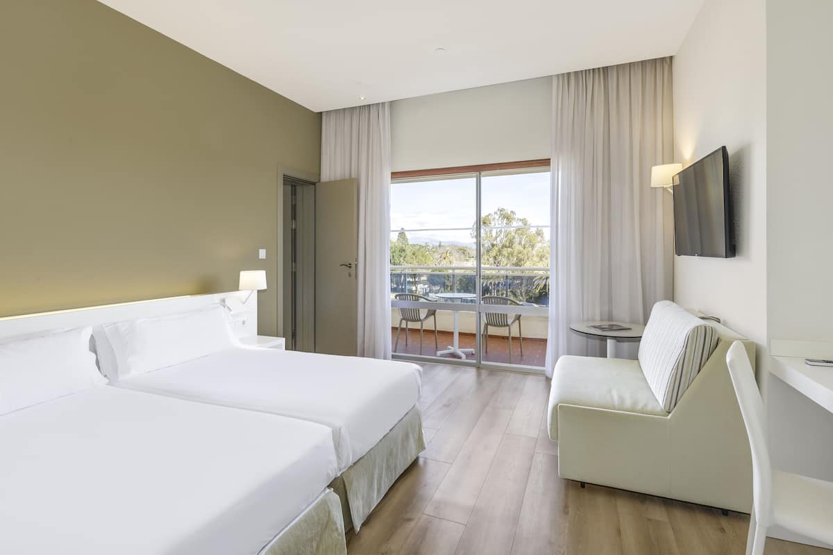 Habitación de hotel con dos camas, un balcón con mesa y sillas, y un televisor en la pared.