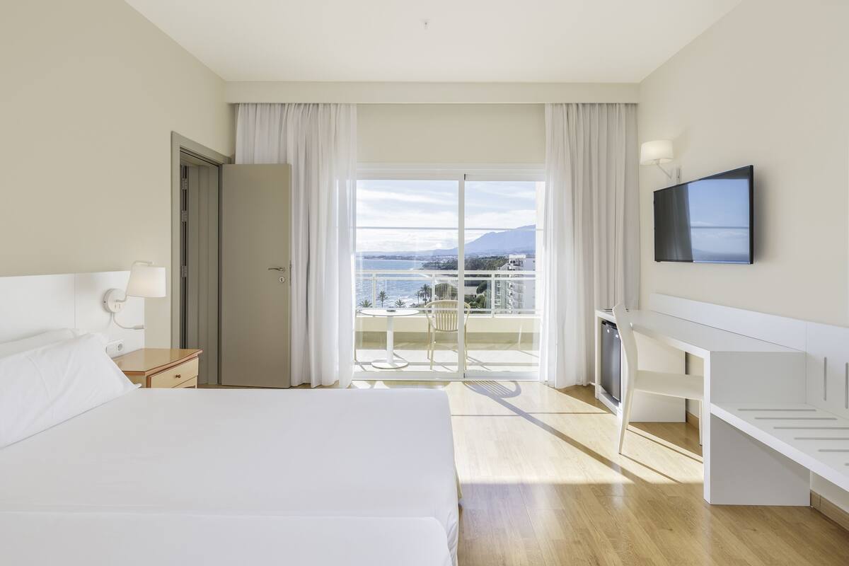 Habitación de hotel con una cama grande, un escritorio, televisión y balcón con vista al mar.