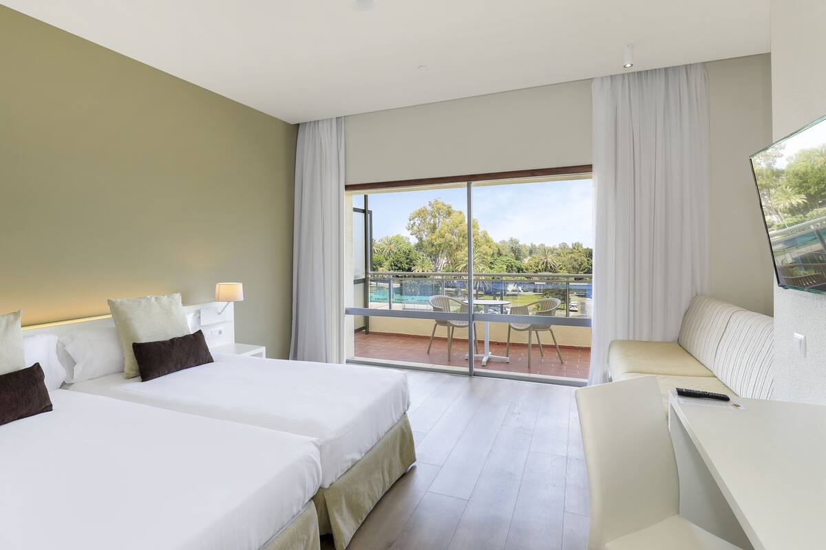Habitación de hotel con dos camas, balcón con vista a la piscina y escritorio con silla.