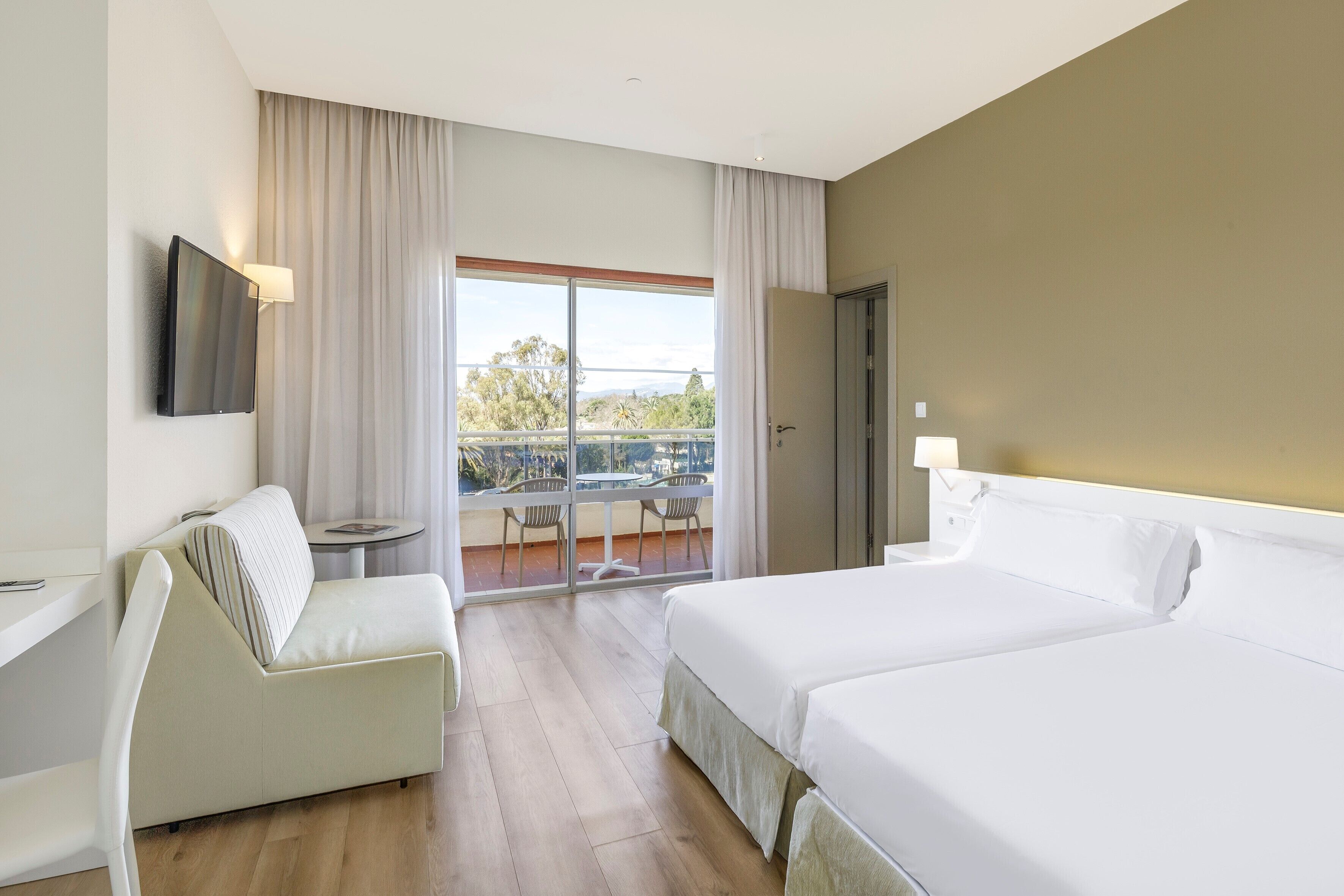 A hotel room with a bed, a sofa, a TV mounted on the wall, and a balcony with a table and chairs.