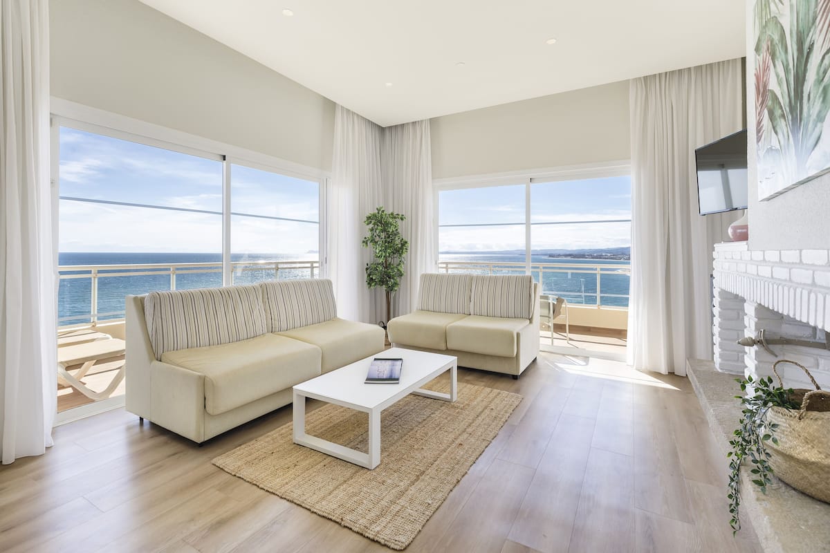Un salón amplio con un ventanal grande con vistas al mar, una chimenea blanca y un moderno conjunto de sofás.