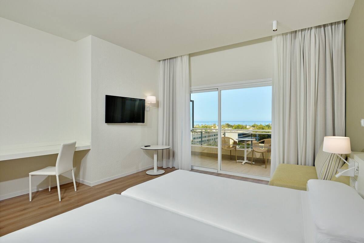 Habitación de hotel con una cama grande, un televisor montado en la pared, una mesita y una silla. Tiene un balcón con vista al océano y edificios.