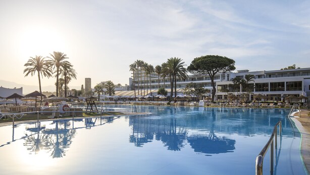 A large outdoor swimming pool with lounge chairs and umbrellas, surrounded by palm trees and modern buildings. - Sol Marbella Estepona - Atalaya Park (Estepona)