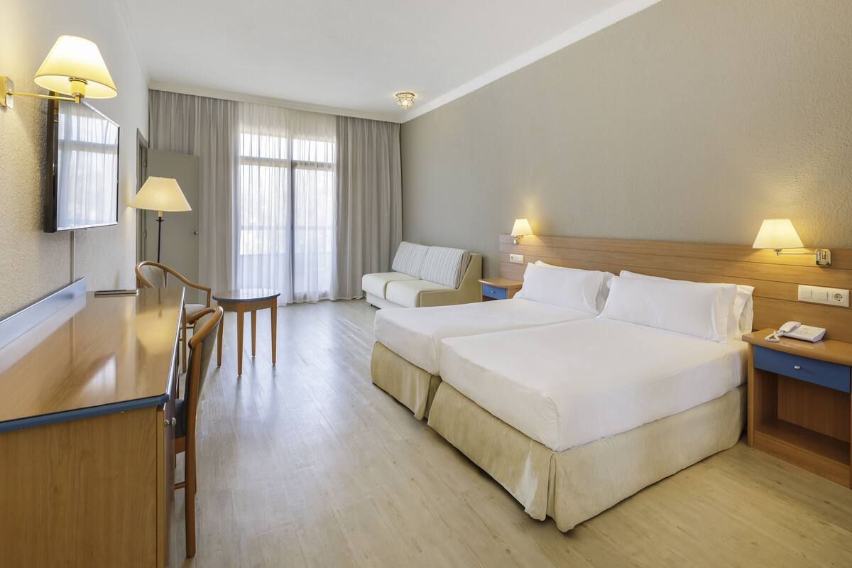 Habitación de hotel con dos camas, un escritorio con sillas, una mesita de noche con lámpara y un ventanal con cortinas.