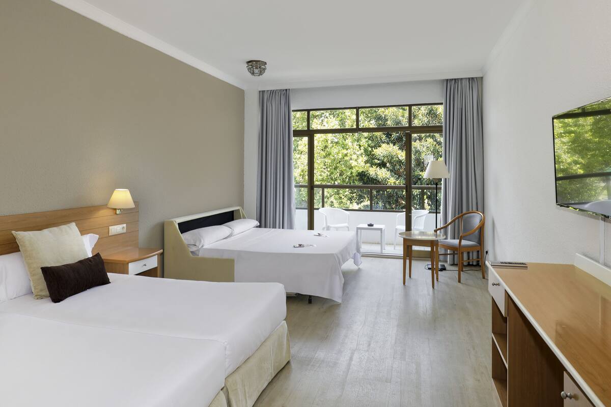 Habitación de hotel con dos camas, un escritorio, una silla, una ventana con cortinas y vistas a árboles.