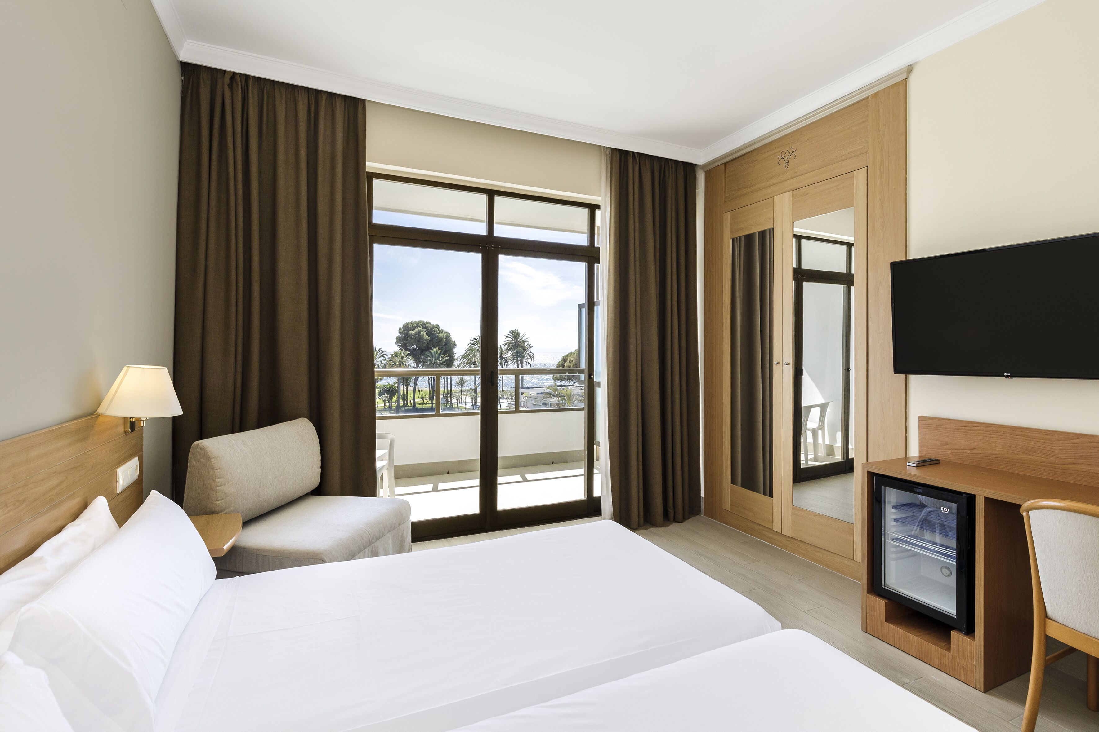 Une chambre d’hôtel dotée d’un grand lit, d’un bureau avec un téléviseur à écran plat, d’une chaise, d’un petit réfrigérateur et d’un balcon avec des rideaux.
