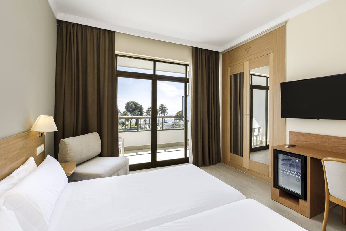 Habitación de hotel con una cama grande, un escritorio con televisor de pantalla plana, una silla, un refrigerador pequeño y un balcón con cortinas.