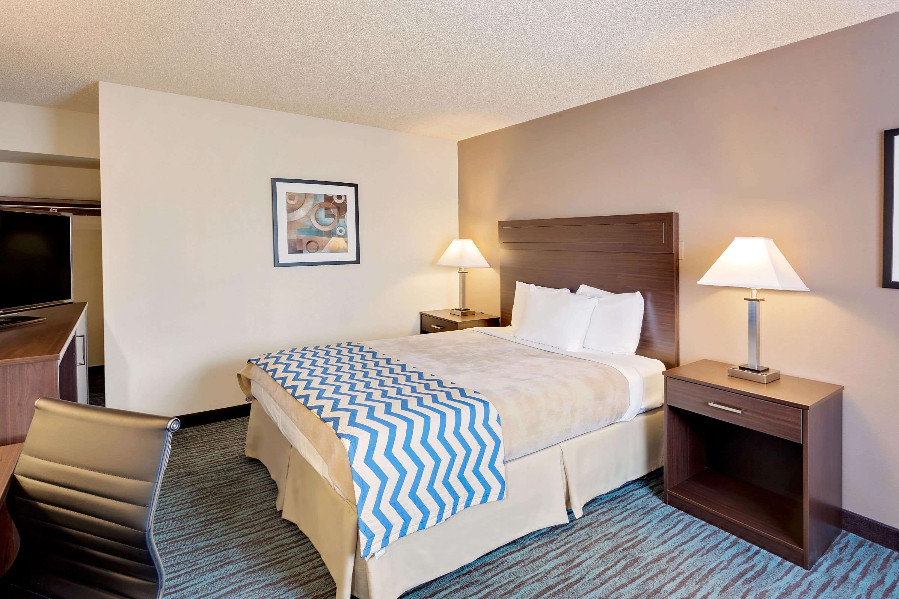 A hotel room with a bed, two bedside tables with lamps, a desk with a chair, and a television.