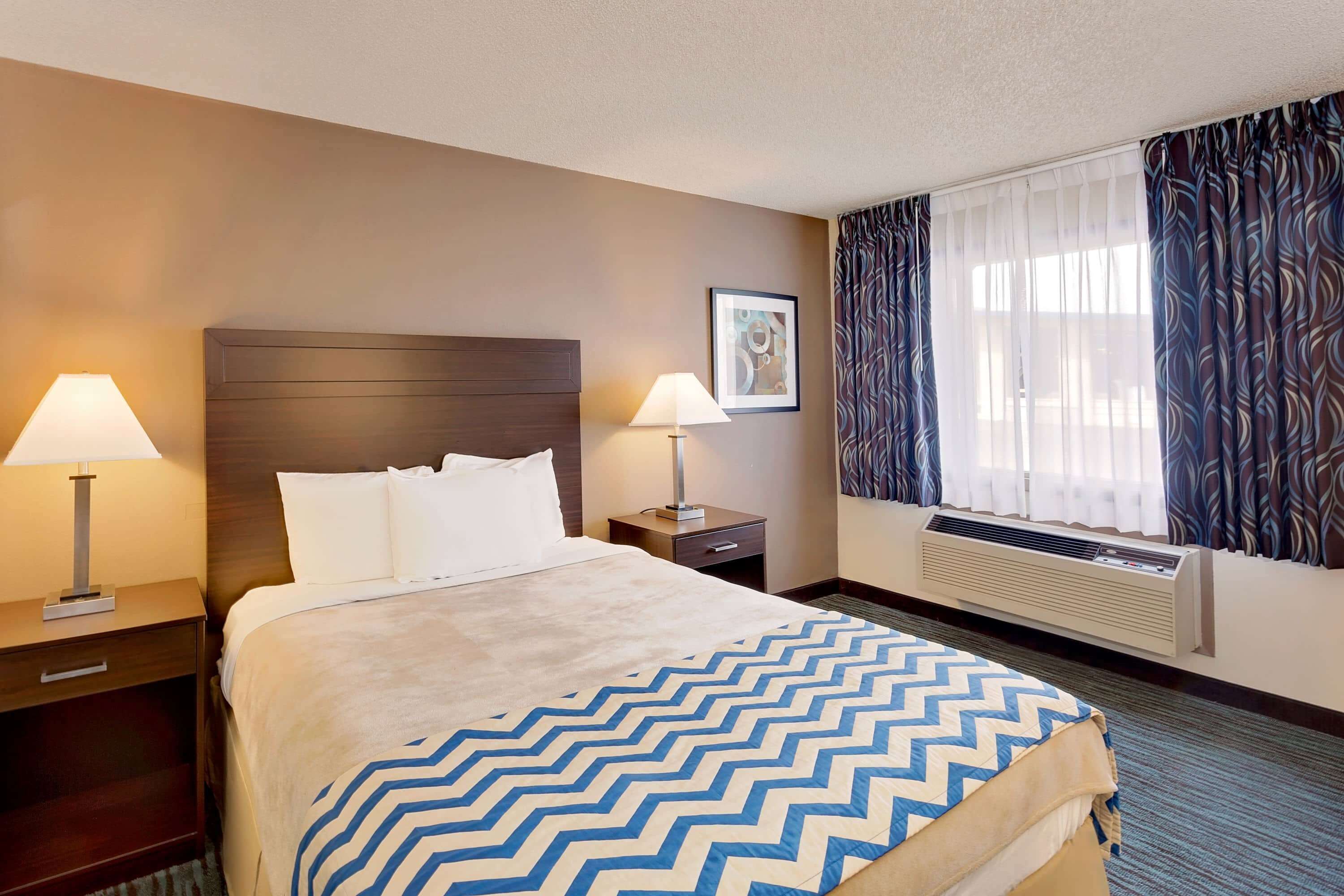 A hotel room with a bed, two bedside tables with lamps, a window with curtains, and a wall-mounted air conditioner.