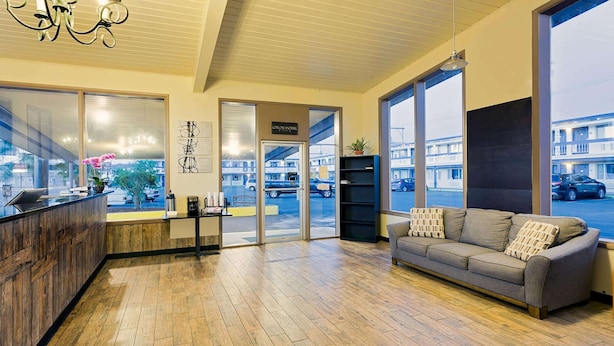 A modern hotel lobby with a wooden floor, a reception desk, a sofa with cushions, and large windows offering a view of the street.
