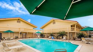 A pool area with lounge chairs and umbrellas, surrounded by a fence and buildings with a red-tiled roof. - La Quinta Inn by Wyndham Lufkin (Lufkin)