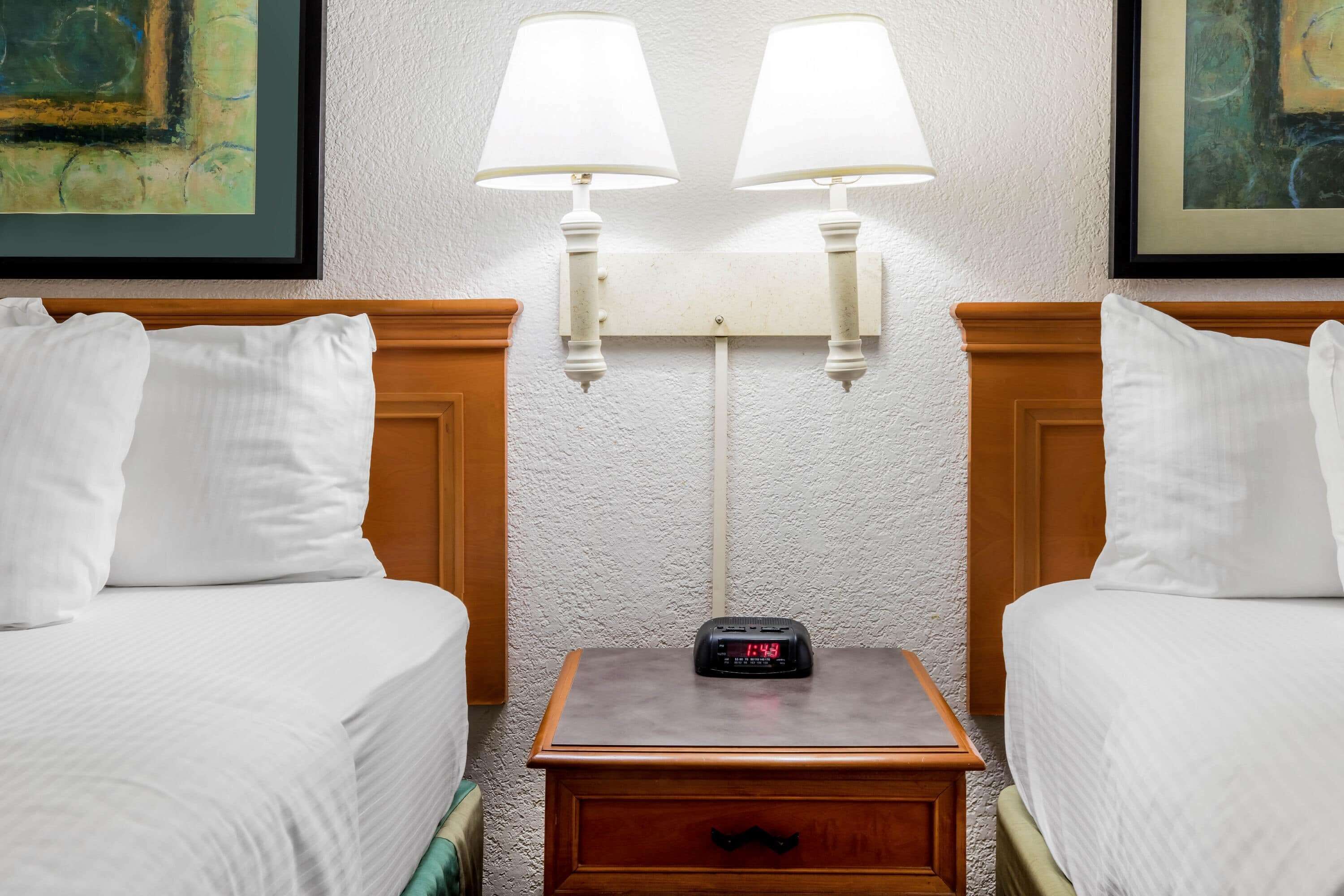 A hotel room with two beds, a nightstand with a clock, and wall-mounted lamps.