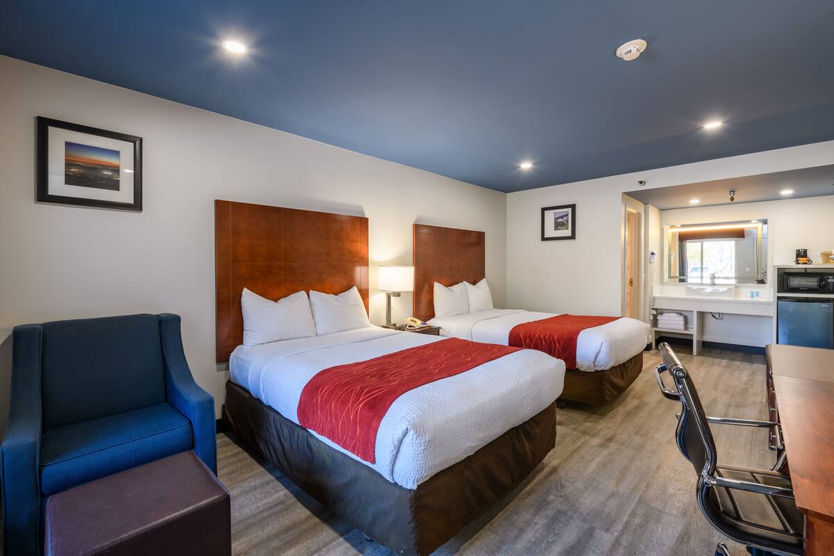 A hotel room with two beds, a desk, a chair, and a small kitchenette.
