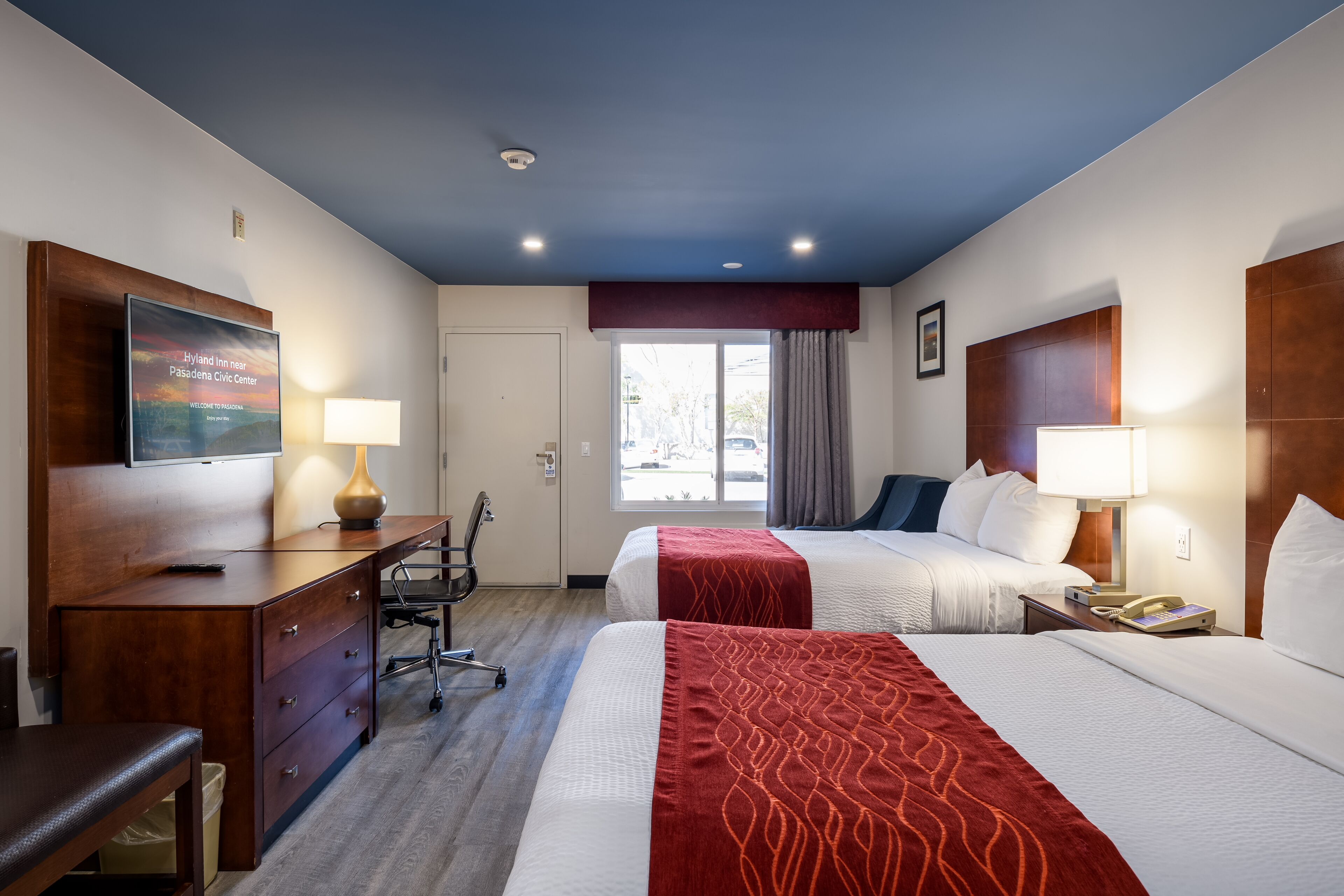 A hotel room with two beds, a desk, a chair, a TV, and a window with curtains.