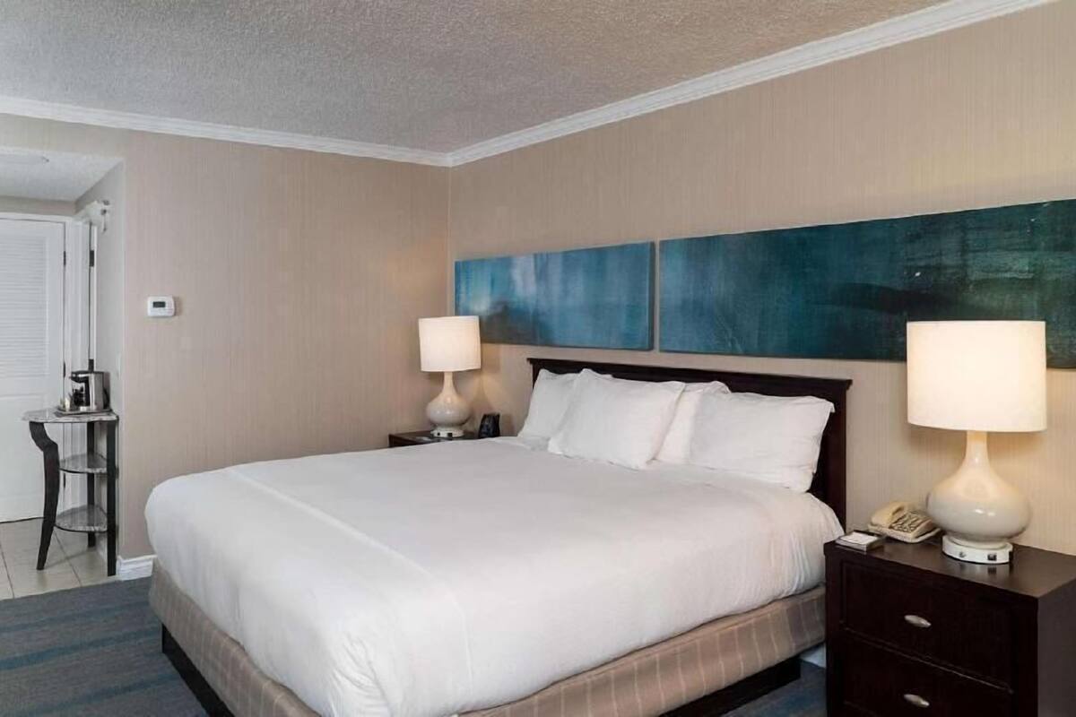 Une chambre d’hôtel avec un grand lit, deux tables de chevet avec des lampes, un petit bureau et une œuvre d’art accrochée au mur.